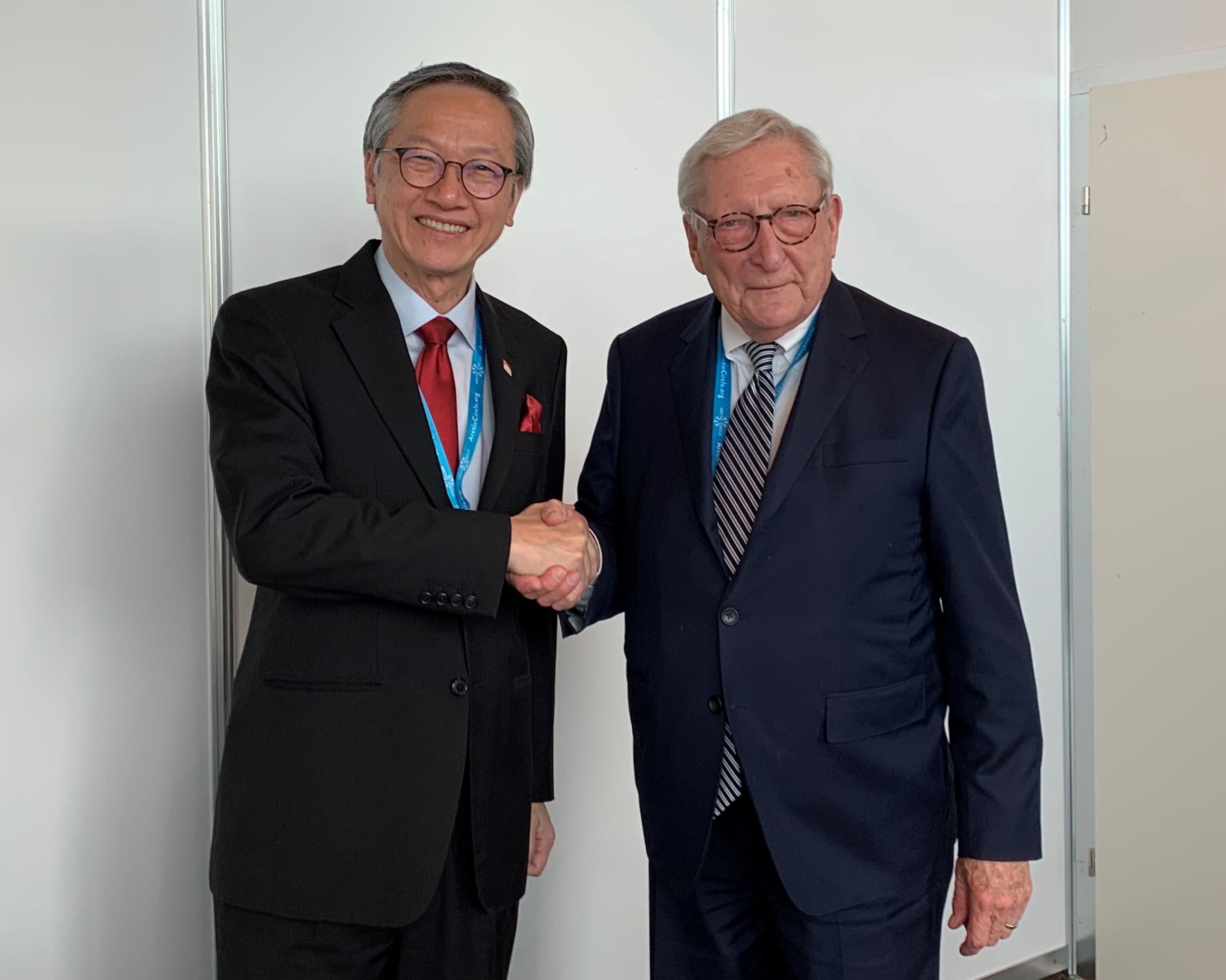 Two men in suits shaking hands; both wear lanyards and glasses.